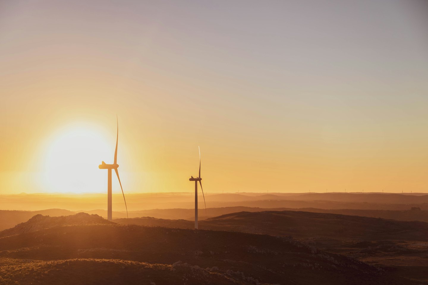 Tuuliturbiinit siluetteina usvaisten kukkuloiden yllä hehkuvaa auringonlaskua vasten.